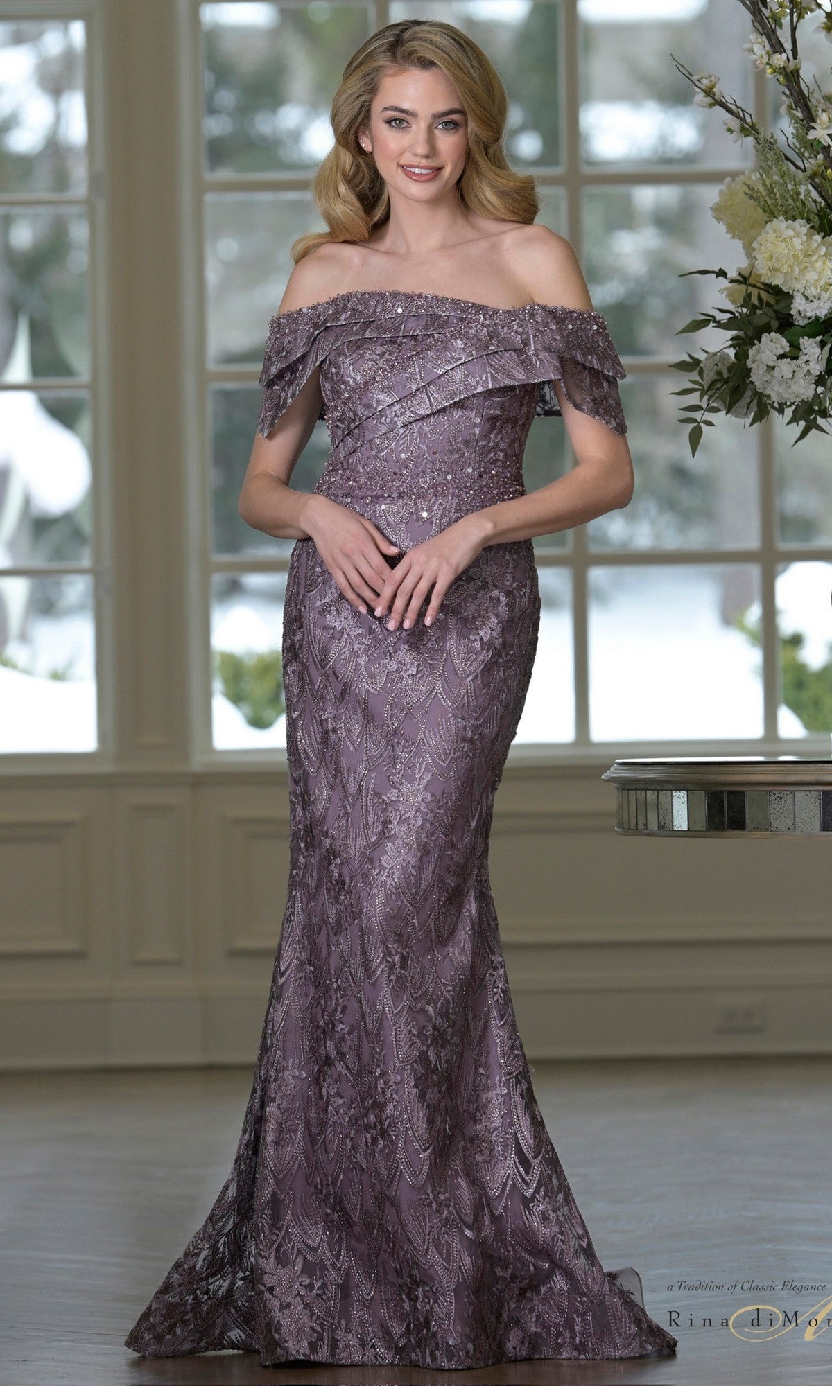 A woman stands indoors wearing the Rina Di Montella RD3531 long formal dress in Victorian Lilac, featuring an off-the-shoulder, floor-length design with lace embroidery and a fitted silhouette—ideal for a mother-of-the-bride.